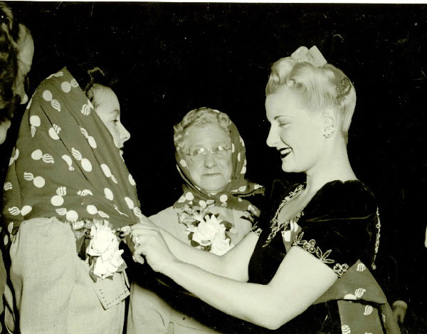 Blonde woman in fancy black dress with glamorous coif ties WOW bandana onto woman worker; a second worker, already wearing her bandana, stands by. 