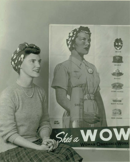  Young woman in WOW bandana seated with 'She's a WOW' poster in background. Poster depicts a blond woman in a WOW bandana and coveralls gazing resolutely into the distance, in foreground is shell casing. the right side of the poster displays the headgear worn by women in other branches of service: WAAC, WAVE etc. 